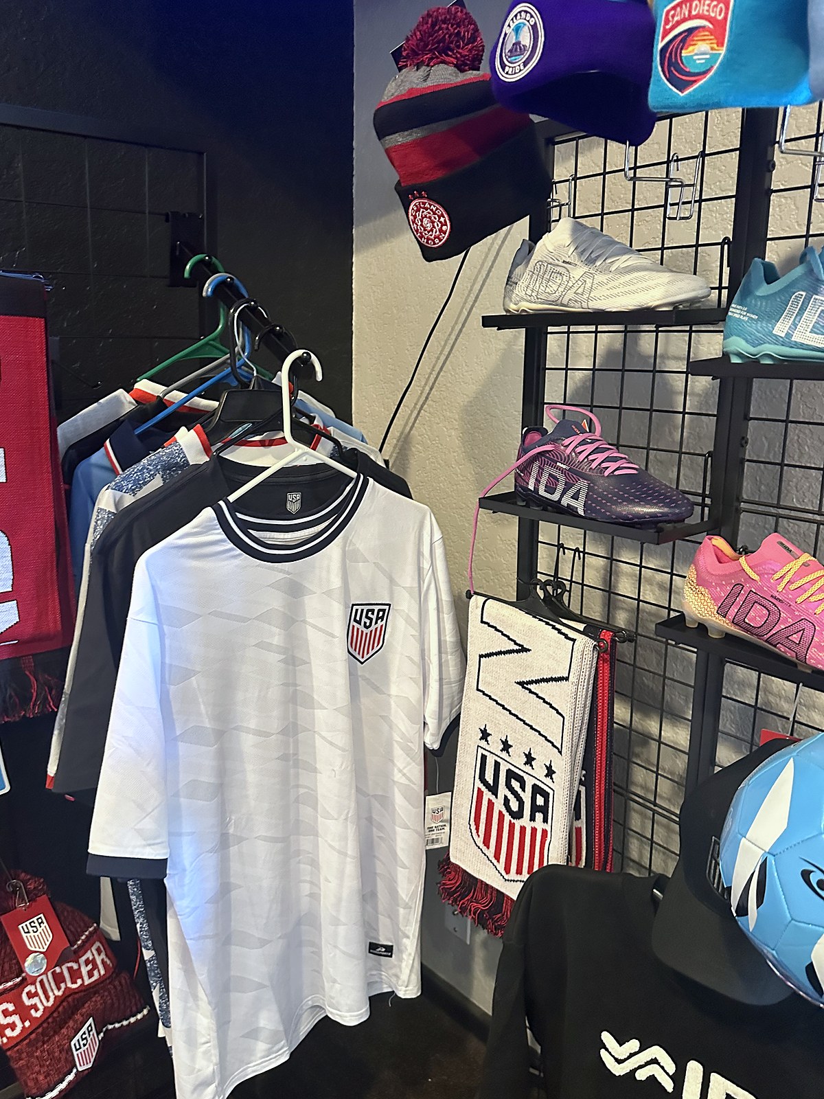 IDA Sports cleats on the display wall at the AMH Goods Pro Shop, with USA jerseys, scarves and NWSL beanies
