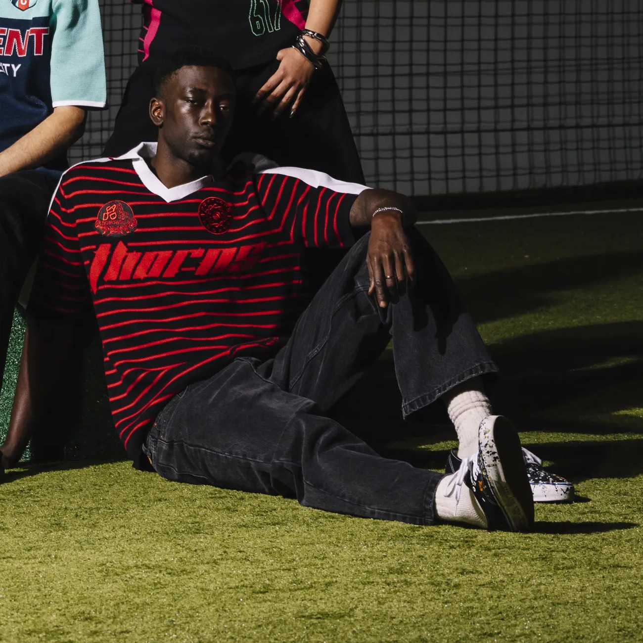 Model wearing the Portland Thorns FC x Dead Dirt NWSL knit polo in black with red stripes, seated on turf